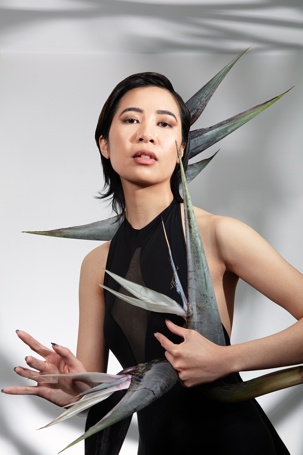 Xuan with smokey eye makeup posed with two bird of paradise flowers against a gray background
